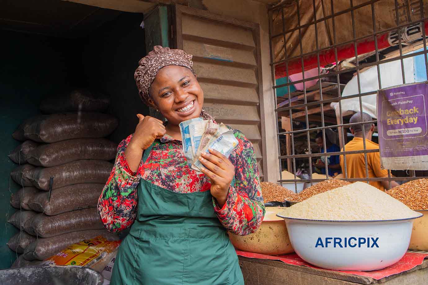 Happy African Woman Holding Naira Note and Giving Thumbs Up in a Bustling Marketplace